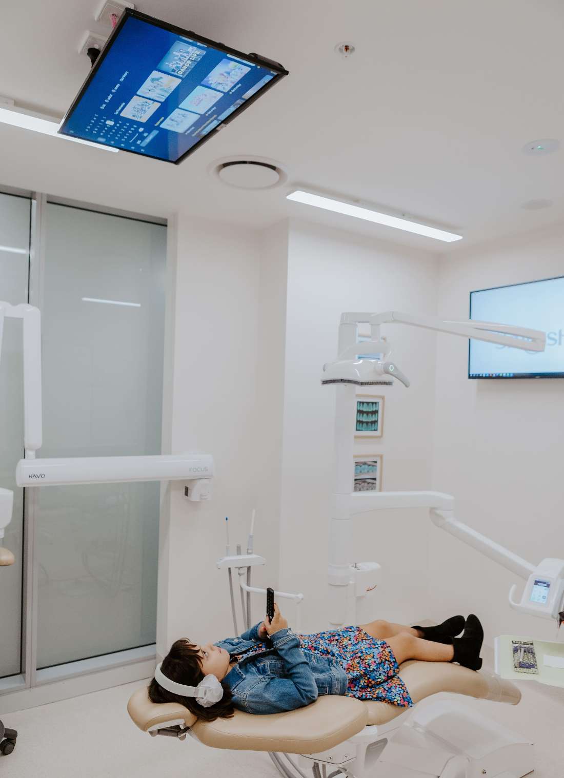 young girl in dental chair changing channels on tv hanging above